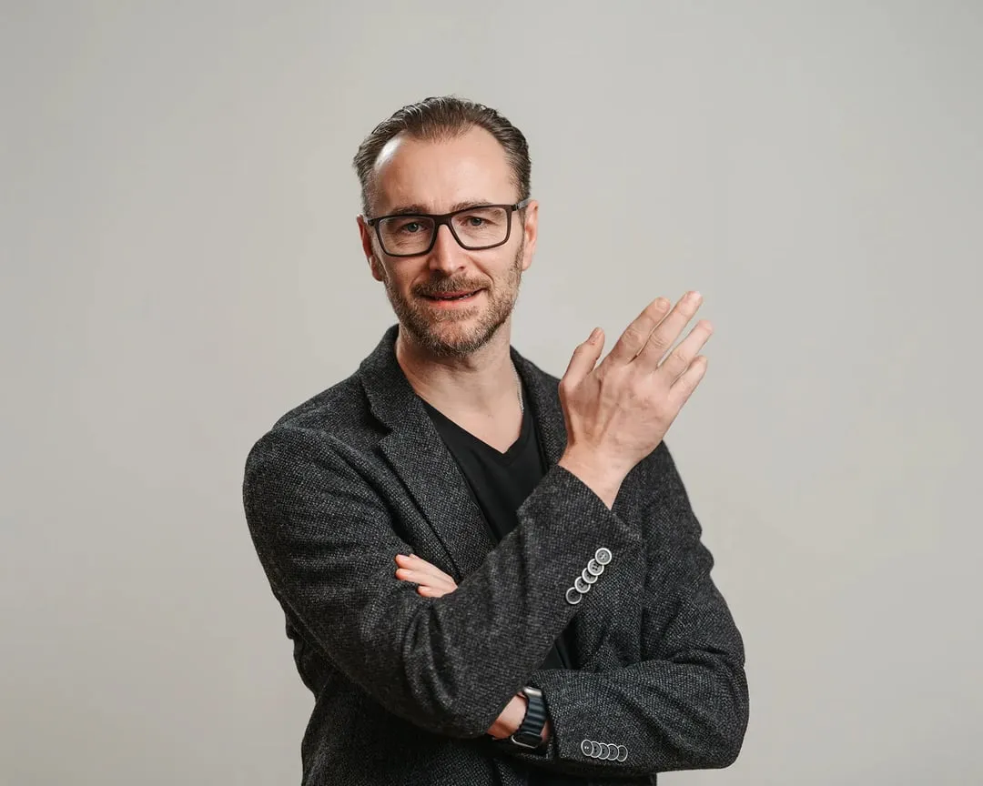 Patrick Lenz mit verschränkten Armen und erhobener Hand vor grauem Hintergrund