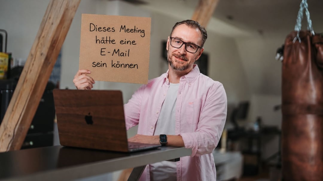 Patrick Lenz am Laptop hält ein Schild mit der Aufschrift Dieses Meeting hätte eine E-Mail sein können